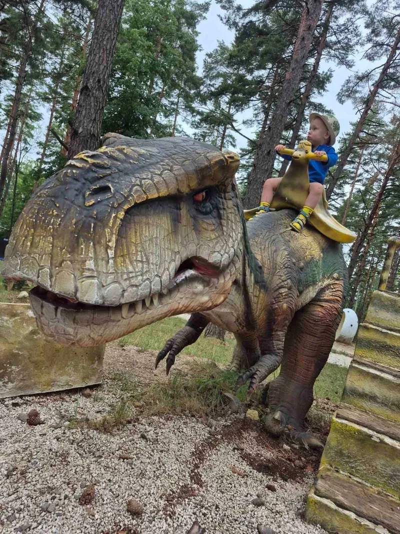 Damián sedí na modelu dinosaura v lese